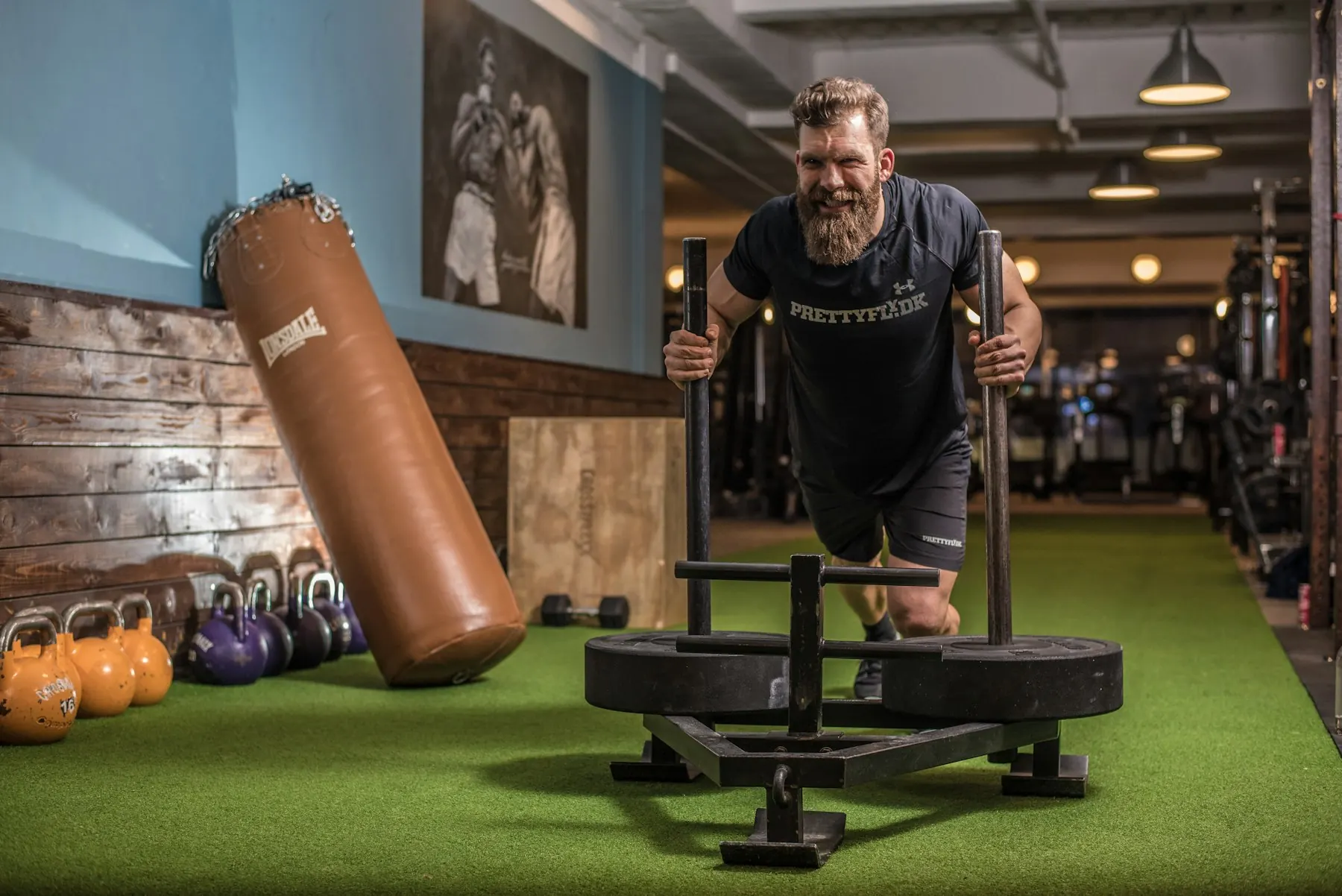 Member pushing a weighted sled across the turf with kettlebells and a heavy bag behind